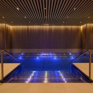 Una moderna piscina cubierta con iluminación azul, detalles metálicos, techo de paneles de madera y escalones a ambos lados crea un ambiente sereno en el spa del Hotel Seventy. Relájese aquí después de disfrutar del spa y masaje con aceites veganos y aromaterapia. - ForTwo - Planes para parejas