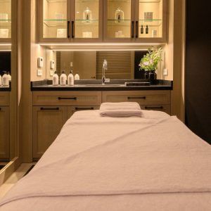 Una camilla de masajes en la sala de spa del Hotel Seventy (Barcelona), con poca luz, está rodeada de un ambiente relajante con lavabo, armarios y plantas. Los aceites veganos realzan este tranquilo refugio con un toque ecológico. - ForTwo - Planes para parejas