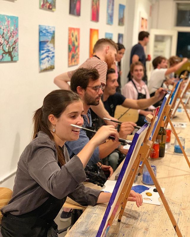En un luminoso estudio de Madrid, la gente participa alegremente en la "Clase de pintura con vino y tapita en Arte Bar", pintando lienzos adornados con impresionantes obras de arte, mientras los suministros de pintura se encuentran esparcidos sobre la mesa. - ForTwo - Planes para parejas