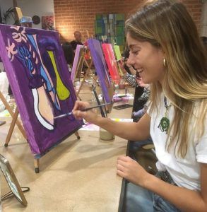En el Arte Bar de Barcelona, una mujer disfruta de una clase de pintura llamada "Clase de pintura con tu copita de vino", mientras pinta un jarrón abstracto morado y verde sobre lienzo. - ForTwo - Planes para parejas