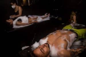 En un spa poco iluminado de Madrid, dos parejas se entregan al Baño turco para parejas en Madrid (Hammam Vital Experience), disfrutando de un relajante masaje de burbujas mientras un terapeuta enmascarado los atiende. - ForTwo - Planes para parejas