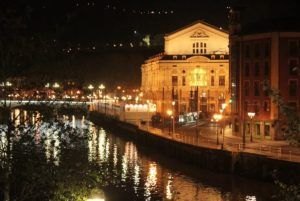 Una vista nocturna de un teatro iluminado junto al río en Bilbao, rodeado de luces de la ciudad reflejándose en el agua, te invita a unirte al Tour gratuito de los misterios y leyendas (Bilbao) y descubrir historias cautivadoras escondidas en sus vibrantes calles. - ForTwo - Planes para parejas