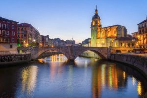 Al atardecer, el puente de piedra de Bilbao se arquea elegantemente sobre el río, flanqueado por edificios iluminados y la torre de una iglesia. El encanto de esta escena es perfecto para el Tour Gratuito de los Misterios y Leyendas (Bilbao), donde los susurros de leyendas y misterios flotan en el aire. - ForTwo - Planes para parejas