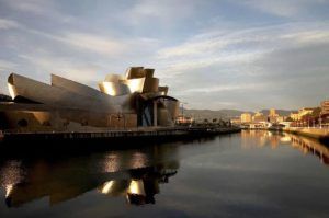 El Museo Guggenheim Bilbao, con su arquitectura metálica reflectante, se alza majestuoso junto a un río tranquilo. Bajo un cielo nublado, crea una vista encantadora que se aprecia mejor durante un tranquilo "Paseo en barco por Bilbao" junto al agua. - ForTwo - Planes para parejas