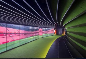Ingresa al pasillo curvo que cuenta con pisos de un verde vibrante y una pared de pantallas iluminadas que muestran el Estadio San Mamés en Bilbao, brindando una vista previa encantadora de tu Tour por el estadio San Mamés. - ForTwo - Planes para parejas