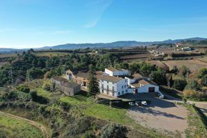 Vista aérea de un paisaje rural con grandes edificios como la Bodega Miquel Jané, rodeado de campos, árboles y colinas lejanas bajo un cielo despejado, un escenario perfecto para la Visita a bodega con degustación y cata de vinos. - ForTwo - Planes para parejas