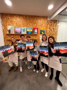 Seis personas con delantales a rayas sostienen pinturas del atardecer, sonriendo a la cámara en Wine Gogh - Pintar mientras bebes vino (Barcelona), capturando sus espíritus creativos en este momento vibrante. - ForTwo - Planes para parejas