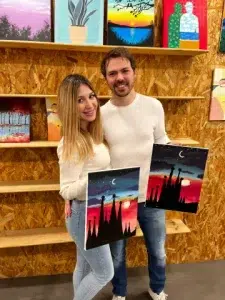 Una pareja sonriente sostiene coloridas pinturas del horizonte de la ciudad en Wine Gogh - Pintar mientras bebes vino (Barcelona), un animado estudio de arte. - ForTwo - Planes para parejas