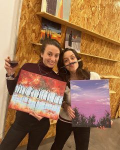 En medio de un entorno artístico, dos personas exhiben con orgullo sus pinturas, mientras una de ellas bebe vino, lo que recuerda a un evento de Wine Gogh - Pintar mientras bebes vino (Barcelona) en la bulliciosa escena artística. - ForTwo - Planes para parejas