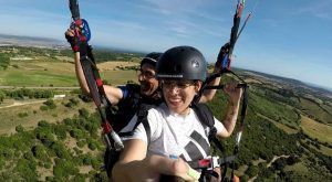 Dos personas disfrutan de un vuelo en parapente biplaza en Vejer (Cádiz) sobre los exuberantes campos verdes y las ondulantes colinas cerca de Cádiz. Hace sol, y ambos llevan casco y sonríen ante la aventura. - ForTwo - Planes para parejas
