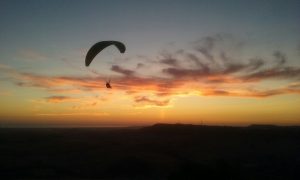 La silueta de un parapente, disfrutando de un "Vuelo en parapente biplaza en Vejer (Cádiz)", se recorta contra un cielo vibrante al atardecer con nubes sobre los impresionantes paisajes de Vejer. - ForTwo - Planes para parejas
