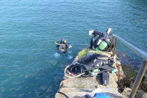 En las tranquilas aguas de Cantabria, un buceador explora mientras otro descansa junto a una barandilla metálica en un borde rocoso, con el equipo a su lado: un entorno perfecto para una experiencia de Bautismo de buceo en Castro Urdiales. - ForTwo - Planes para parejas