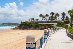 Disfrute de un día relajante en una playa soleada con pasarela y barandillas azules, con palmeras y vistas lejanas al océano. Perfecto para disfrutar del encantador Paseo en Barco en Santander. - ForTwo - Planes para parejas