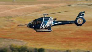 Un helicóptero azul y blanco se desliza con gracia sobre un paisaje, dejando al descubierto campos y vegetación, que recuerda a un pintoresco Vuelo en helicóptero por la Costa del Sol (Marbella). - ForTwo - Planes para parejas