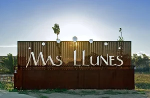 Un cartel metálico con “MAS LLUNES” marca la entrada a la Bodega Mas Llunes en Garriguella, Girona, donde el sol se pone detrás y las plantas la rodean. - ForTwo - Planes para parejas
