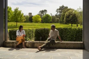 Dos personas se relajan en la cornisa de una terraza del Hotel Món, abrazadas por la exuberante vegetación y los árboles en un día soleado, disfrutando de una serena experiencia que recuerda a una "experiencia medieval" cerca del histórico monasterio de St. Benet. - ForTwo - Planes para parejas