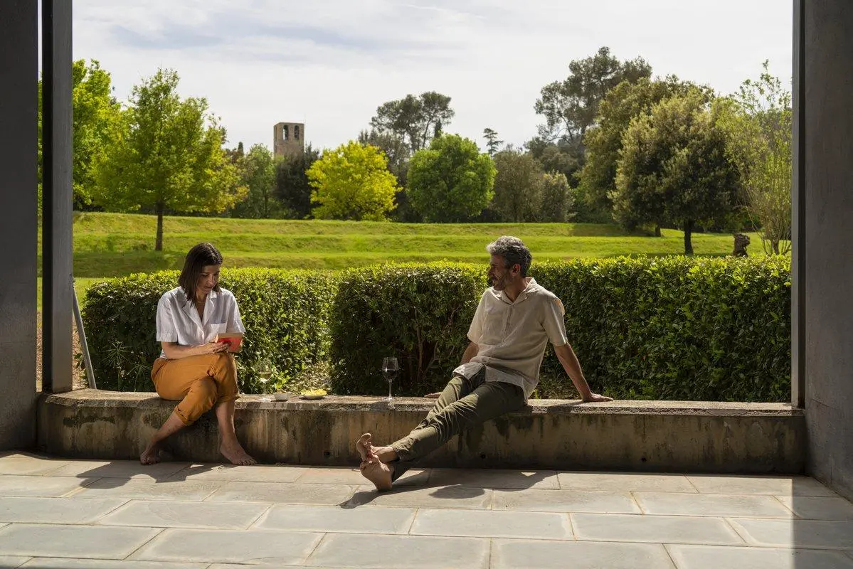 Dos personas se relajan en la cornisa de una terraza del Hotel Món, abrazadas por la exuberante vegetación y los árboles en un día soleado, disfrutando de una serena experiencia que recuerda a una "experiencia medieval" cerca del histórico monasterio de St. Benet. - ForTwo - Planes para parejas