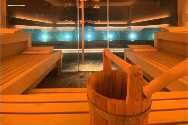 El "Circuito de bienestar en Wellington Hotel & Spa (Madrid)" incluye una acogedora sauna con bancos de madera y un cubo, que ofrece una vista perfecta de la piscina iluminada a través de una pared de cristal, creando una experiencia de oasis de lujo. - ForTwo - Planes para parejas