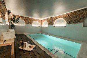 Disfrute de la lujosa piscina cubierta del Hospes Puerta de Alcalá en Madrid, con Spa Privado con Comida o Cena, rodeada de elegantes arcos de ladrillo y agua iluminada. Disfrute de champán y postre en la terraza de madera, sumiéndose en una serena relajación. - ForTwo - Planes para parejas