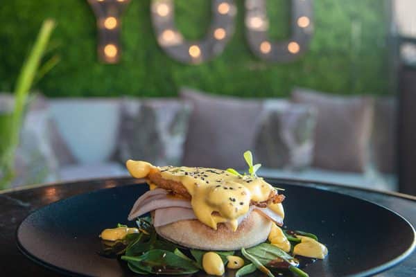 Huevos Benedict con salsa holandesa servidos en un plato negro frente a una exuberante vegetación en el Hotel Only YOU Atocha de Madrid. Disfrute de este plato durante el Brunch de Fin de Semana: Saborea el Delicioso Sabor del Descanso. - ForTwo - Planes para parejas