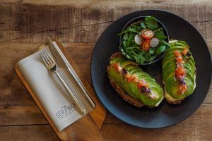 Dos tostadas de aguacate con aderezos y una ensalada pequeña en un plato negro, con cubiertos sobre una tabla de madera. Disfruta de este "Brunch de Fin de Semana: Saborea el Delicioso Sabor del Descanso" en Only YOU Hotel Atocha (Madrid). - ForTwo - Planes para parejas