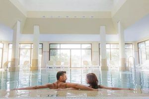 Una pareja disfruta del Ritual de relajación acuática con masaje de esencias y copa de cava en pareja en Playa Granada Club Resort & Spa, relajándose en una piscina cubierta con grandes ventanales que enmarcan el sereno telón de fondo. - ForTwo - Planes para parejas