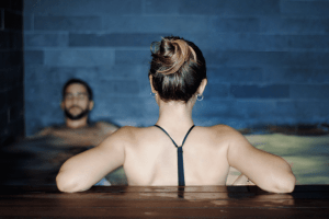 Una persona con un moño frente al jacuzzi y otra relajándose en el agua, contra una pared de azulejos oscuros, creando un ambiente sereno donde la indulgencia se encuentra con la tranquilidad en Desayuno Buffet, Circuito Spa y Masaje en pareja en Serena Spa, Meliá Barcelona Sky. - ForTwo - Planes para parejas