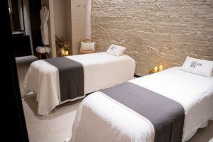 En una habitación serena con paredes de ladrillo e iluminación de velas en Yurbban Passage Hotel & Spa, dos mesas de masaje con mantelería blanca le esperan para ofrecerle el "Masaje vegano con aromaterapia y acceso al spa en Yurbban Passage Hotel & Spa (Barcelona)" para una experiencia rejuvenecedora. - ForTwo - Planes para parejas