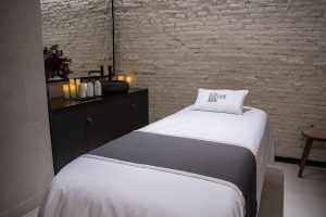 Disfrute de un masaje vegano con aromaterapia en el Yurbban Passage Hotel & Spa, Barcelona, en una serena sala de spa con mesa de masaje, sábanas blancas, velas y un armario negro contra una pared de ladrillos. - ForTwo - Planes para parejas