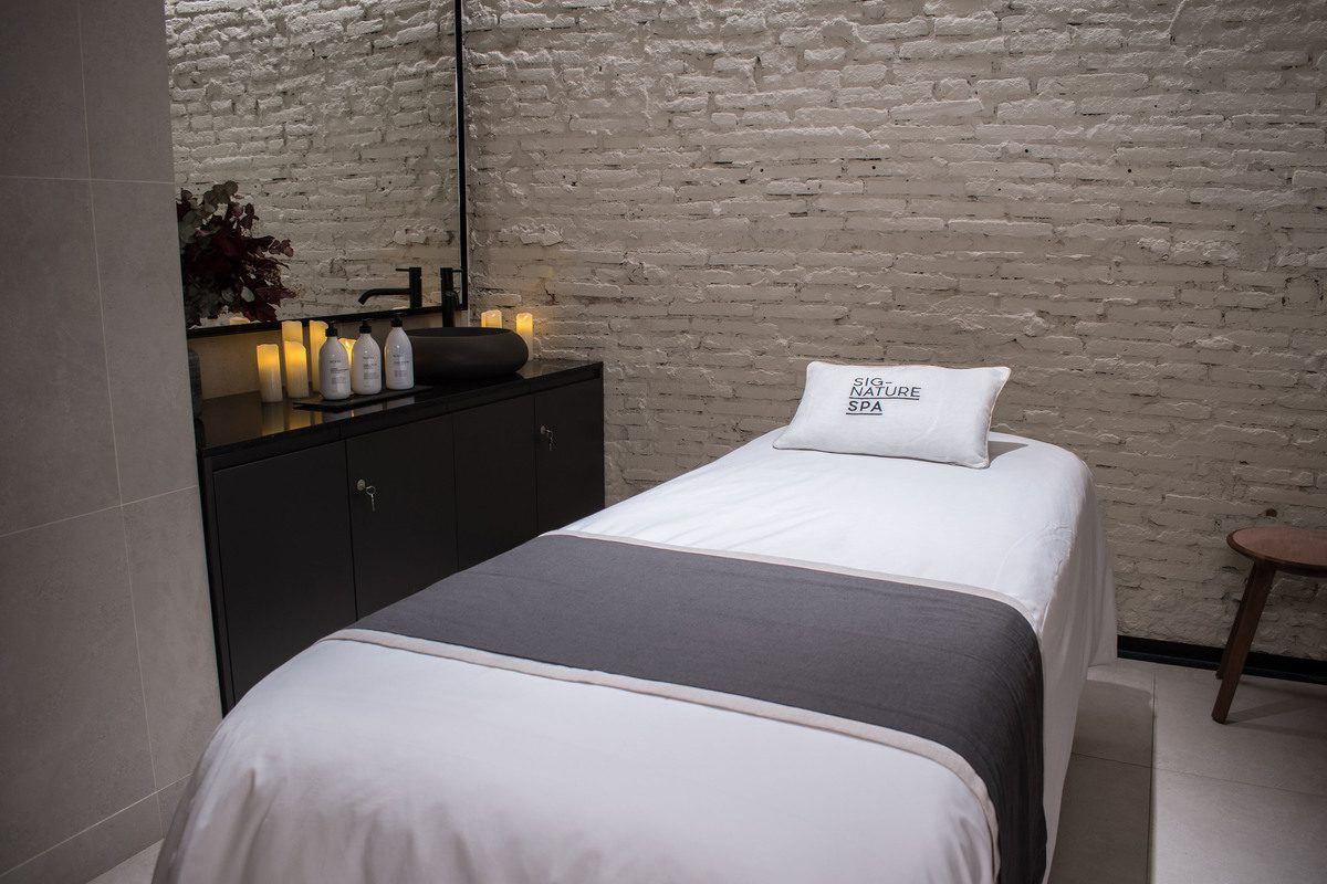 Una sala de spa minimalista en Barcelona con una cama de masaje, velas encendidas y productos de spa contra una pared de ladrillos ofrece el escenario ideal para un "Masaje en pareja con técnicas de relajación profunda" en Yurbban Passage Hotel & Spa. - ForTwo - Planes para parejas
