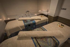 La sala de spa poco iluminada del Hotel Catalonia, Granada cuenta con dos mesas de masaje con toallas enrolladas, caminos de mesa estampados y velas, creando un ambiente sereno para disfrutar del "Circuito de aguas, cava y masaje de esencias para parejas". - ForTwo - Planes para parejas