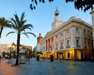 Únete al Free tour por Cádiz para explorar un edificio histórico con una icónica torre de reloj, ubicado entre palmeras ondulantes en una encantadora plaza al atardecer, y descubrir sus cautivadoras historias. - ForTwo - Planes para parejas