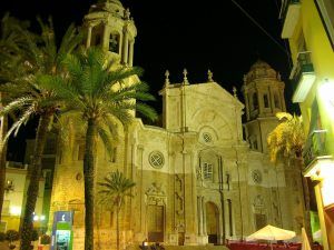 Vista nocturna de una catedral iluminada con torres gemelas en Cádiz, rodeada de palmeras, capturando la esencia de antiguas leyendas y misterios atemporales explorados en la "Visita guiada de los misterios y leyendas de Cádiz. - ForTwo - Planes para parejas