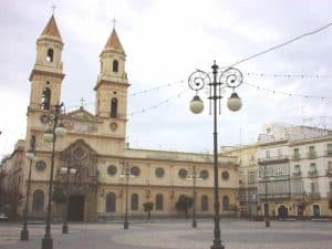En una plaza vacía, una histórica iglesia gaditana con dos campanarios se yergue entre farolas y edificios bajo un cielo nublado. El aire se impregna del encanto de la "Visita guiada de los misterios y leyendas de Cádiz", como si las propias paredes susurraran historias antiguas. - ForTwo - Planes para parejas