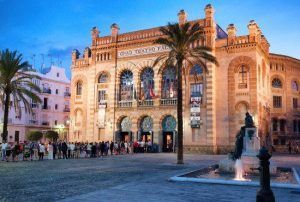 El histórico teatro, resplandeciente al atardecer gaditano, atrae a visitantes entusiastas. Con palmeras ondulantes y una fuente reluciente en las cercanías, el escenario ofrece un preludio sugerente al cautivador "Tour gratuito por Cádiz". - ForTwo - Planes para parejas