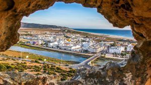A través de una abertura rocosa, Visita por la Costa de Tarifa descubre Zahara de los Atunes con edificios blancos, un río encantador bajo un pintoresco puente y una playa iluminada por el sol que se encuentra con el mar azul: una joya escondida. - ForTwo - Planes para parejas