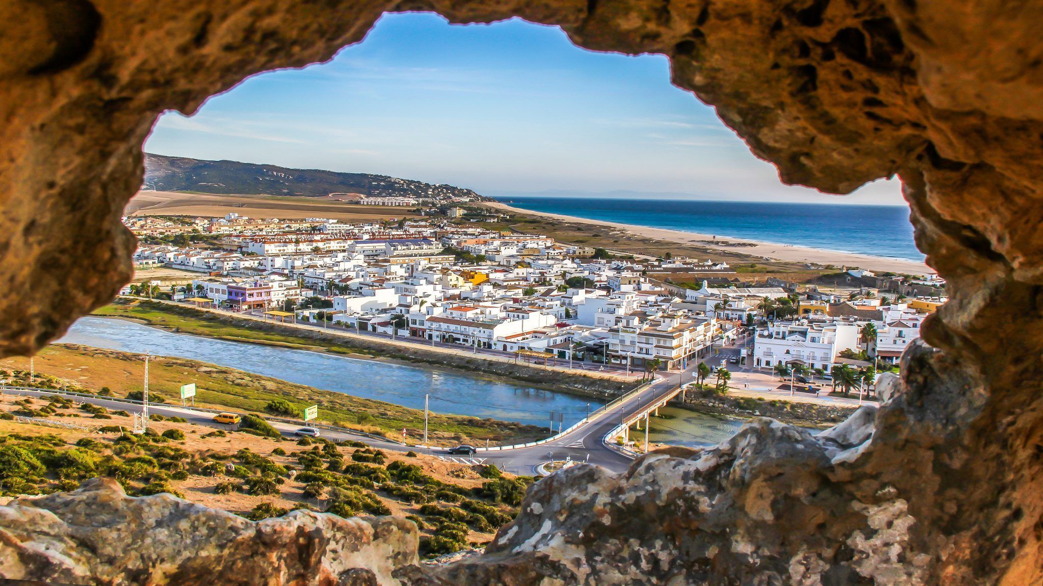 A través de una abertura rocosa, Visita por la Costa de Tarifa descubre Zahara de los Atunes con edificios blancos, un río encantador bajo un pintoresco puente y una playa iluminada por el sol que se encuentra con el mar azul: una joya escondida. - ForTwo - Planes para parejas
