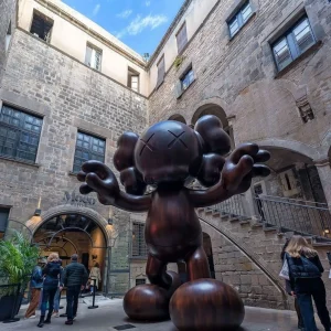 En un patio de piedra, una gran estatua caricaturesca con ojos tachados, que recuerda a una obra de Banksy, está rodeada de gente y arquitectura histórica. Podría encajar perfectamente en las vibrantes calles cercanas al Museo MOCO de Barcelona, conocido por "Banksy y más: Sin colas". - ForTwo - Planes para parejas
