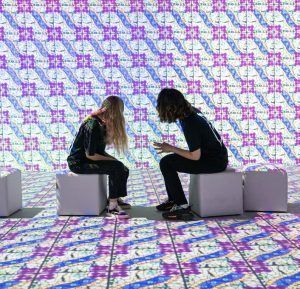 Dos personas se sientan en cubos en una habitación adornada con los coloridos patrones digitales geométricos de "Dragones y Modernismo en Barcelona: Experiencia inmersiva", que se hacen eco del Art Nouveau y de los motivos de dragones. - ForTwo - Planes para parejas