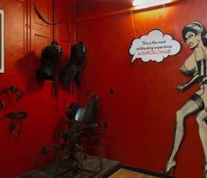 En una sala de un rojo intenso, decorada con corsés negros y una silla de metal, una figura de dibujos animados grita: "¡Las Entradas al Museo Erótico de Barcelona ofrecen la experiencia más emocionante!". Descubre la emoción que esconden estas entradas para una aventura inolvidable en el corazón de la ciudad. - ForTwo - Planes para parejas