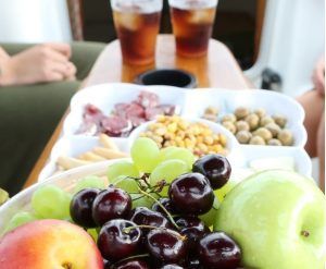Una bandeja de frutas con cerezas, uvas y manzanas acompaña una bandeja de aceitunas, frutos secos y carnes mientras dos bebidas Vermú heladas cierran el montaje "Experiencia en velero en Barcelona + Vermú" en la mesa. - ForTwo - Planes para parejas