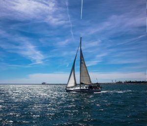 Un velero se desliza elegantemente sobre el mar resplandeciente bajo el cielo azul de Barcelona, un rastro de vapor arriba crea el escenario perfecto para una tranquila Experiencia en velero en Barcelona + Vermú. - ForTwo - Planes para parejas