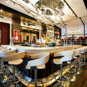 Interior de bar moderno con taburetes beige, un mostrador central lleno de bebidas y un cartel de "Hamburguesa en Hard Rock Café Barcelona" en la pared, que recuerda la atmósfera vibrante que se encuentra en este popular lugar. - ForTwo - Planes para parejas