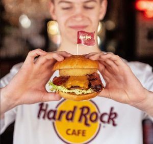 Vistiendo una camiseta de Hard Rock Café, una persona disfruta de la Hamburguesa en Hard Rock Café Barcelona cubierta con lechuga, queso y tocino. - ForTwo - Planes para parejas