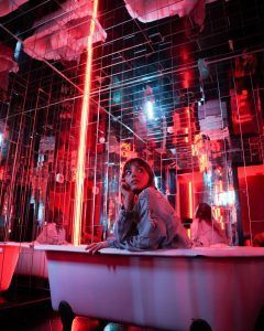 Bañada por el resplandor vibrante de las luces de neón rojas y rodeada de paredes espejadas, una mujer se sienta en una bañera en IKONO Barcelona - La experiencia inmersiva del 2023, experimentando un escape trascendente hacia un caleidoscopio de reflejos y tonos. - ForTwo - Planes para parejas