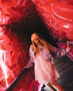 Una mujer con un vestido blanco y una chaqueta vaquera camina por un túnel de color rosa vibrante, mirando hacia arriba, como si estuviera entrando en IKONO Barcelona - La experiencia inmersiva del 2023. - ForTwo - Planes para parejas
