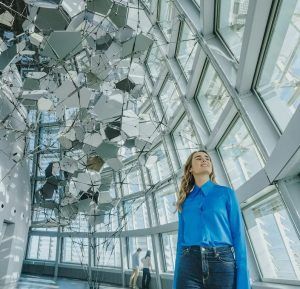 Una mujer con una camisa azul se encuentra en un moderno edificio de cristal, con una escultura geométrica encima que ofrece vistas impresionantes similares a las del Mirador Torre Glorias. - ForTwo - Planes para parejas