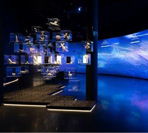 La exhibición del museo, similar al Mirador Torre Glorias, muestra elementos suspendidos en cajas de vidrio en medio de paredes futuristas iluminadas de azul y un piso reflectante que brilla con elegancia moderna. - ForTwo - Planes para parejas
