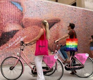 Dos personas en bicicletas rosas con bolsas de colores vibrantes se toman un descanso durante el Barcelona Street Art Tour cerca de un gran mural de mosaico de una cara sonriente en la colorida Barcelona. - ForTwo - Planes para parejas