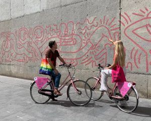 En las vibrantes calles de Barcelona, dos ciclistas con bolsas de arcoíris se detienen junto a una pared de grafitis con figuras rojas abstractas, que encarnan la esencia del arte callejero local y capturan el espíritu del Barcelona StreetArt Tour. - ForTwo - Planes para parejas